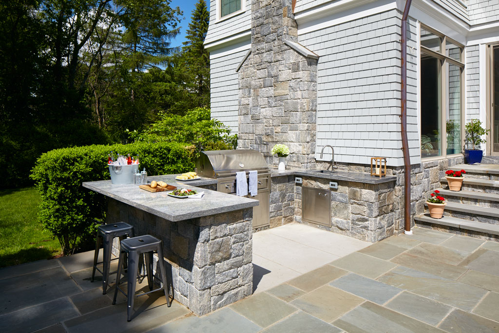 Outdoor stone kitchen with built-in stainless steel grill, sink, granite countertops, bar seating, and decorative potted plants
